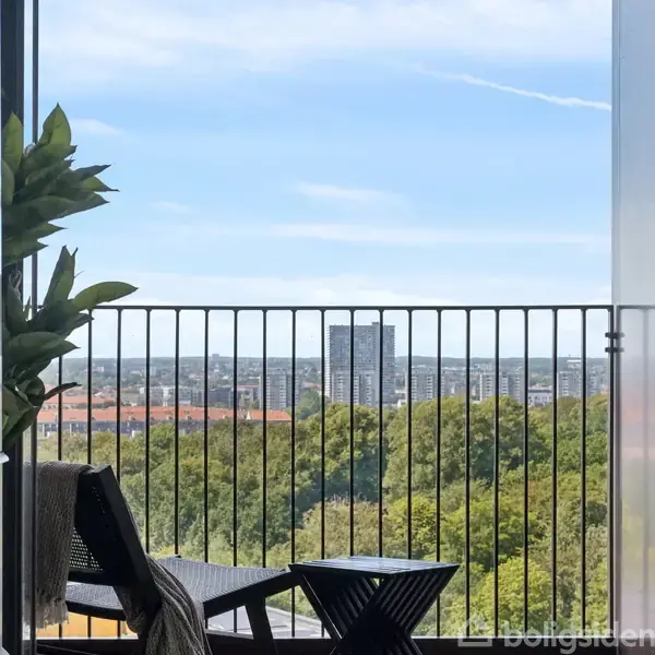En balkon med sort rækværk står foran en udsigt over en by med højhuse, grønne træer og blå himmel. Venstre side har en grøn plante ved rækværket.