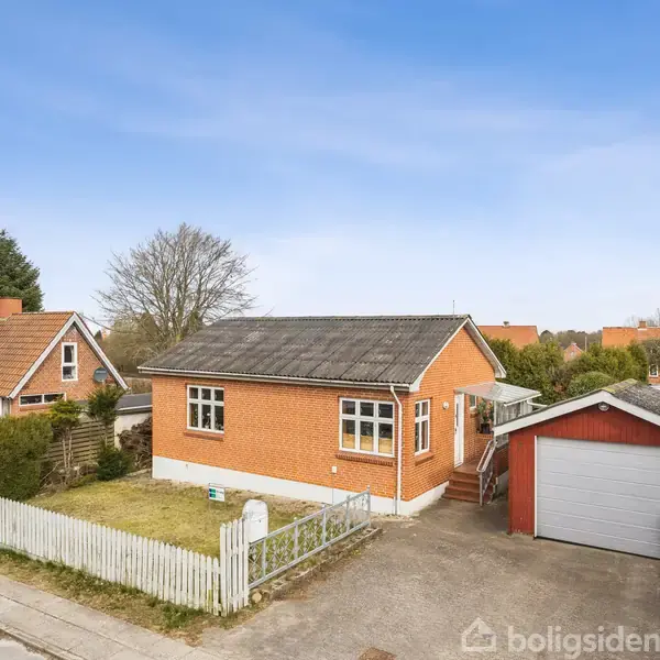 Et rødt murstenshus med hvide vinduer står ved siden af en hvid malet stakit, foran en garage. Det er placeret i et villakvarter med græsplæne og træer.