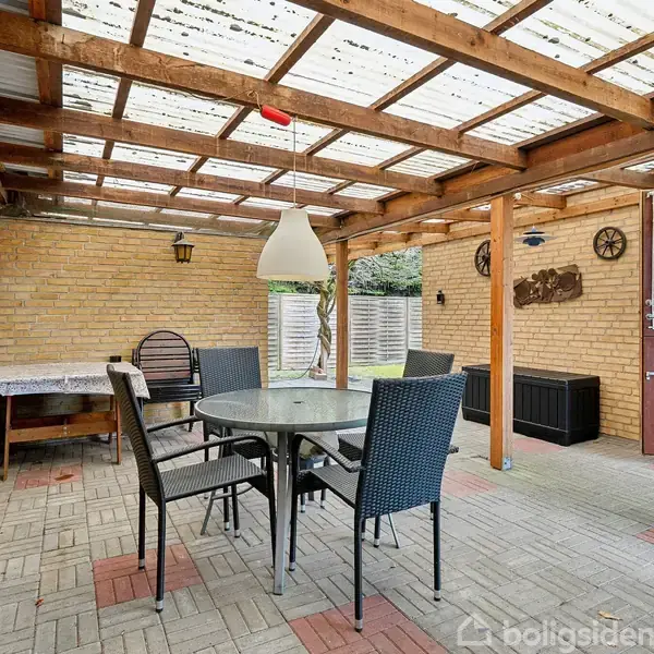 Rundt havebord med sorte stole står under en overdækket terrasse. Murstensvægge omgiver det, og en glaspaneldør åbner til haven. Små pyntegenstande hænger på væggene.