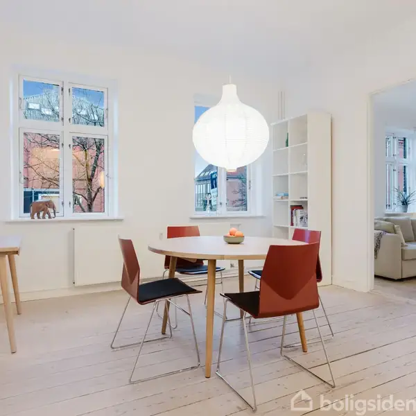 Rundt spisebord med fire stole står under en stor, hvid pendellampe i et lyst rum. Vindue og hylde ses i baggrunden, med en stue til højre.