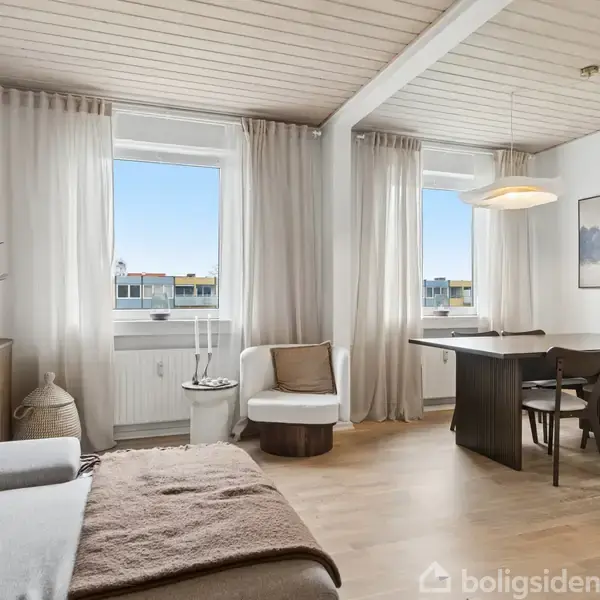 Stue med sofa og bord, sollys skinner gennem hvide gardiner på vinduer, træloft, vægdekorationer samt spisebord med fire stole. To runde puder ligger på sofaen.