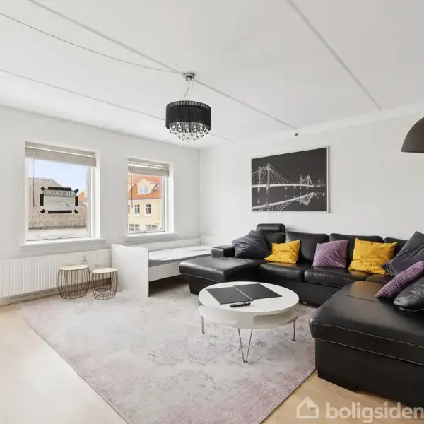 Sort hjørnesofa med farverige puder står på tæppe i en lys stue. Tre vinduer slipper naturligt lys ind. Et kunstværk hænger på væggen over sofaen.