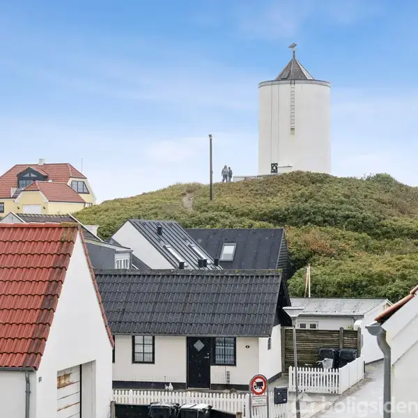 Et hvidt tårn står på en bakke dækket af grøn vegetation. I forgrunden ses huse med røde tegltage i en bymæssig setting under en blå himmel.