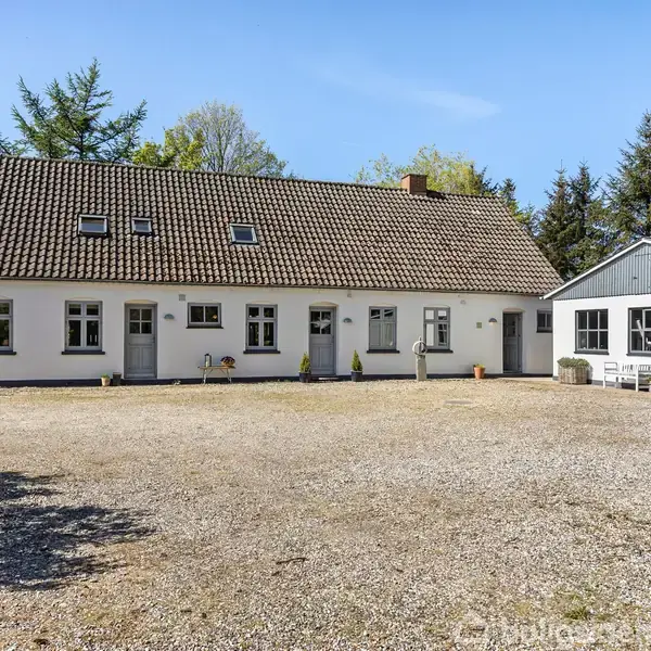 En hvid landlig bygning med rødt tegltag står stille i en gårdsplads omgivet af beplantning og skov. Ingen synlig tekst.