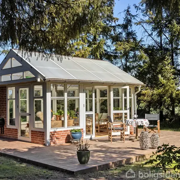 Et drivhus af glas og mursten står på en terrasse, omgivet af træer. Indvendigt er der planter og møbler, hvilket skaber en hyggelig og afslappende atmosfære i haven.