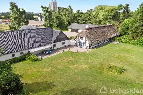 Gårdhus med stråtag, placeret i en grøn have. Omkringliggende bygninger omfatter skure og træer, der giver en landlig atmosfære.
