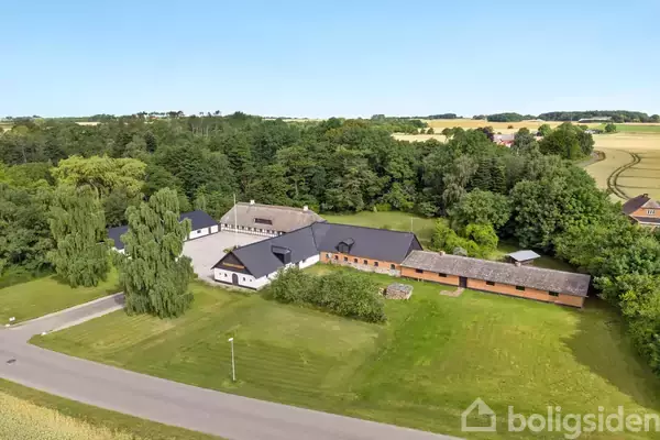 En gårdbygning med sort tag ligger i et frodigt, grønt område omgivet af træer og en åben mark. En vej løber langs den ene side af ejendommen.