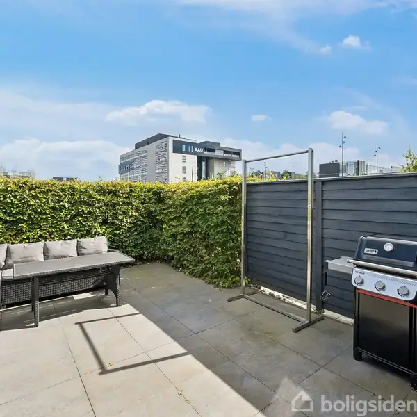En terrasse med en grå hjørnesofa og et bord foran en grøn hæk. Til højre står en grill ved et sort træhegn. I baggrunden ses moderne bygninger.