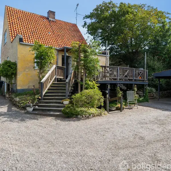 Gult hus med rødt tegltag, smukke trapper til verandaen, omgivet af grønne buske. Grusvej foran, carport til højre med en bil, træer i baggrunden.