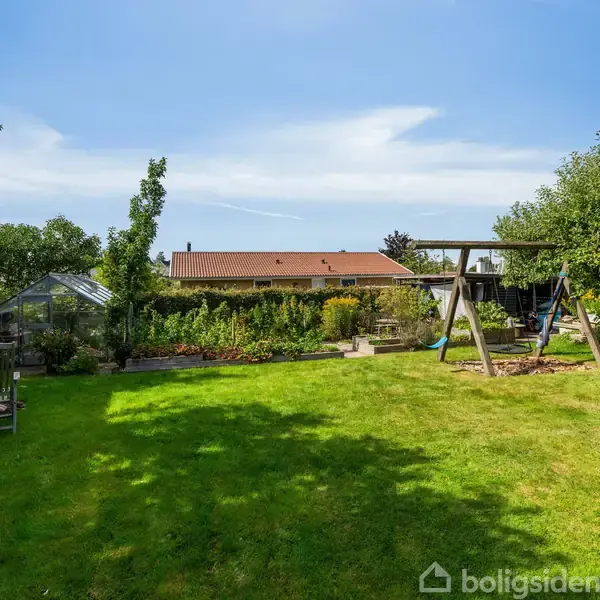 En gynge står i en grøn have, hvor der også er en hvilestol til venstre og buske rundt om. I baggrunden ses et hus med rødt tag.