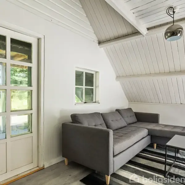 Grå sofa står i hjørnet af et lyst rum med skrå loft. Ved siden af er et lille vindue og en glasdør. Gulvtæppe og sofabord fuldender hyggen.