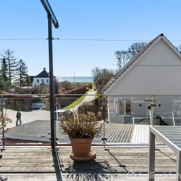 Terrasse med trepotter, der står stille, omgivet af et glasrækværk. I baggrunden ses en vej, huse, træer og havudsigt under en klar blå himmel.