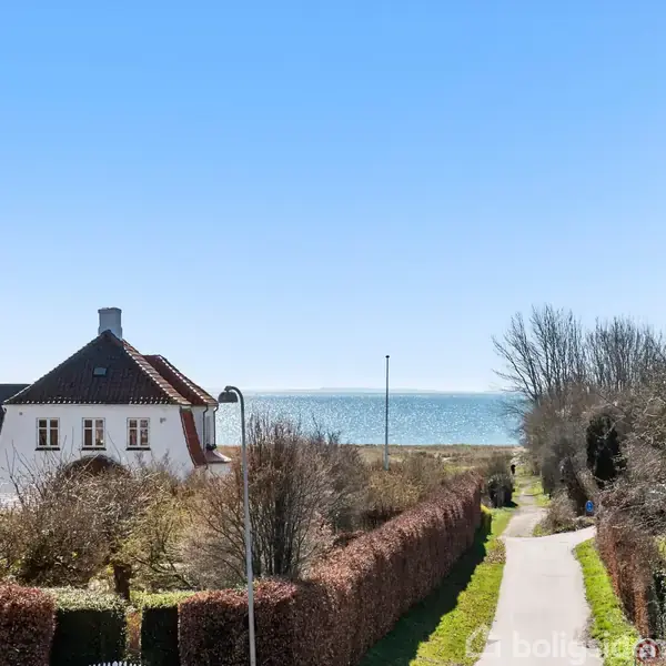 Hus med rødt tag står stille omgivet af have og buske, beliggende ved en gangsti med udsigt over havet i det fjerne under en klar blå himmel.
