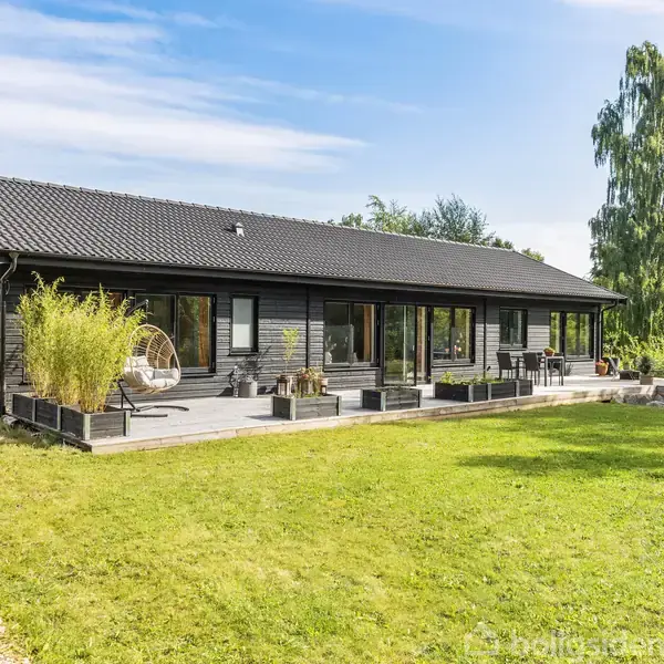Et moderne, sortmalet hus med en bred træterrasse omgivet af plantekasser. Terrassen rummer en hængestol og havemøbler. Huset ligger i et frodigt område med græsplæne og træer.