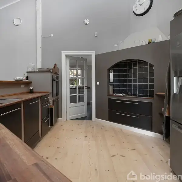 Køkken med sort-skabsfronter og trægulv. Kogeplade indbygget i en sort flise væg. En åben glasdør fører til en anden del af huset.