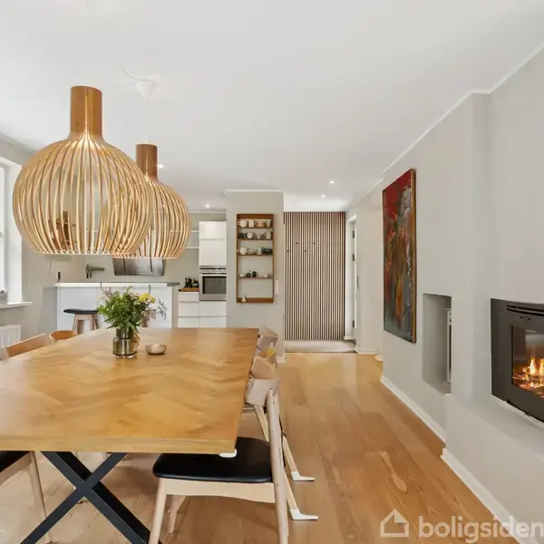 Et træbord med stole er midtpunktet. Det er i et moderne køkken-alrum med store vinduer, trælamper hængende fra loftet og en vægmonteret pejs med ild.