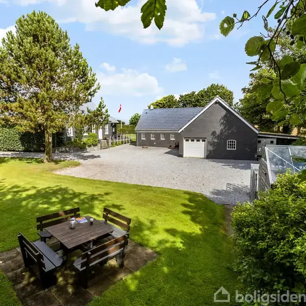 Hus med gråt tag og hvid garage står omgivet af grønne træer og bænkesæt i en velplejet have. Græsplænen dominerer forgrunden, mens en indkørsel ses i baggrunden.