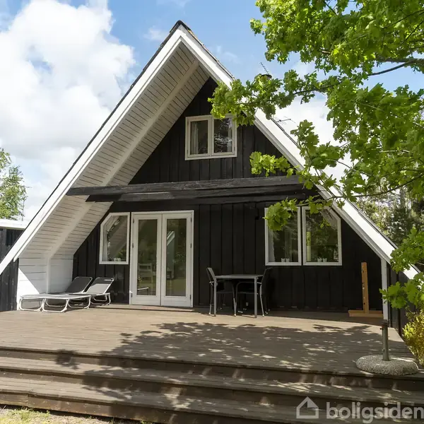 A-formet sort træhus står stille på en åben terrasse med hvide døre og vinduer, omgivet af grønne træer under en blå himmel med hvide skyer.