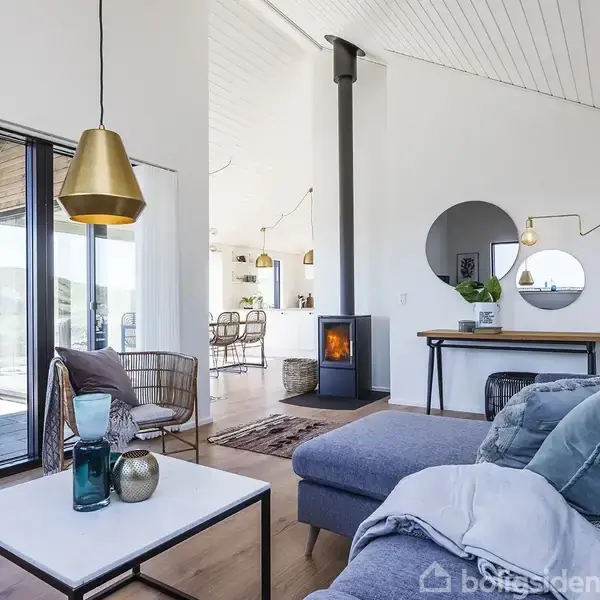 En moderne stue med en grå sofa, beplantede puder og tæpper. En brændeovn står ved væggen, omgivet af et rundt spejl og lamper. Store vinduer åbner mod en terrasse.