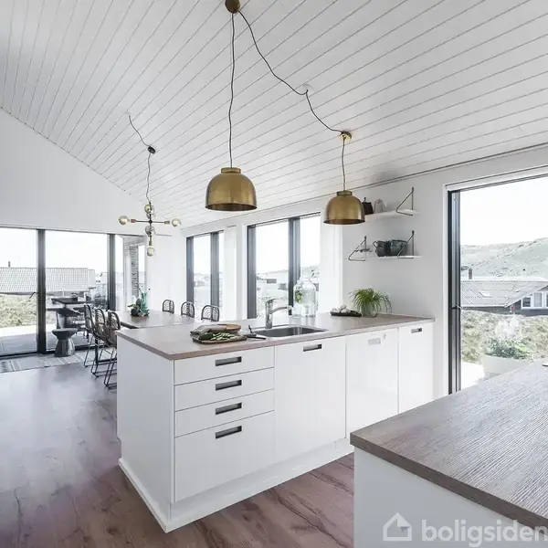 Køkkenet er lyst og moderne med hvide skabe og træbordplader. To messinglamper hænger over køkkenøen. Store vinduer giver udsigt til natur og terrasse udenfor.