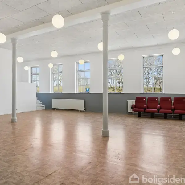 Stor, tom sal med træparketgulv og hvidmalede vægge. Flere runde lamper hænger fra loftet. To søjler deler rummet. Døre og en radiator langs væggene. Ingen tekst.