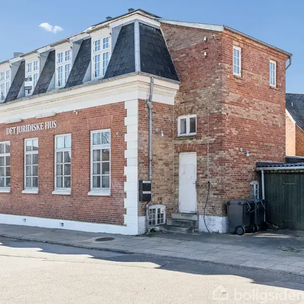Bygning med røde mursten og sort tag. Skilt på væggen siger "DET JURIDISKE HUS". Bygningen ligger på en stille vej med fortov, og der er en garage ved siden af.