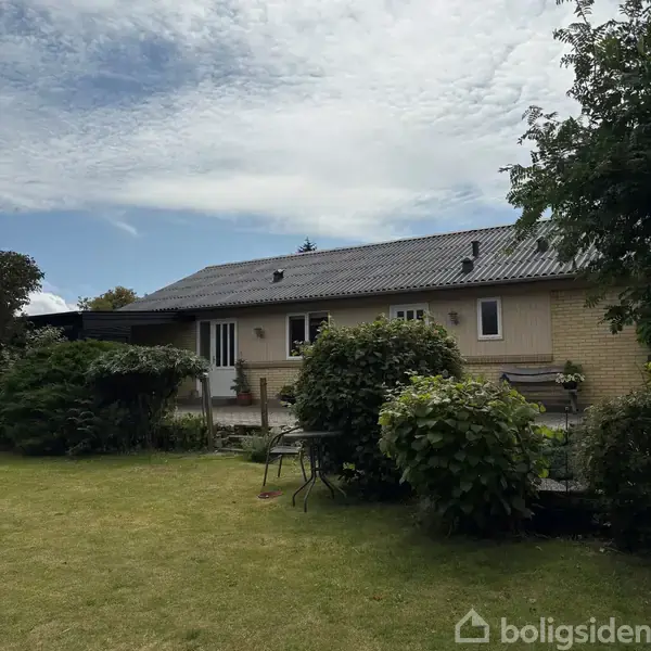 Hus står stille omgivet af en velplejet have med grønne buske og træer. Himlen er overskyet, og der står et bord og stole på græsplænen foran huset.