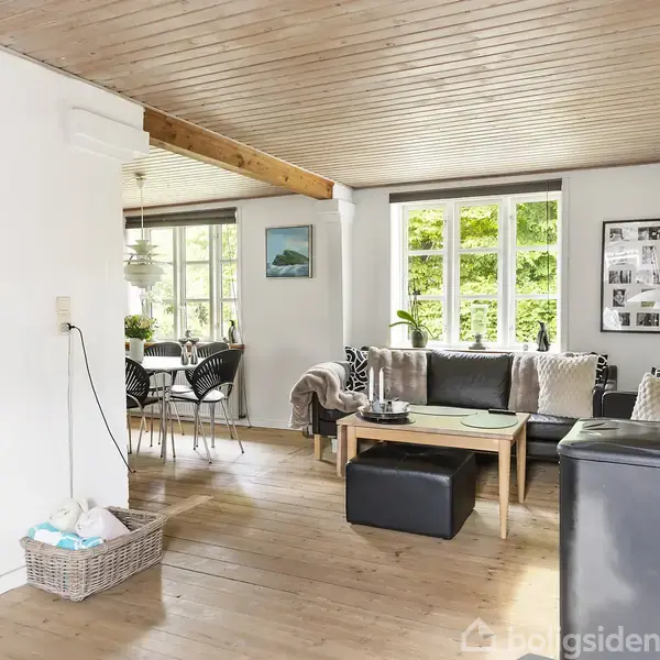 En lys stue med trægulv og træloft. Sofaen står ved vinduet med grøn udsigt. Et spisebord med stole er i baggrunden. Vægdekorationer og en kurv med brænde ses også.