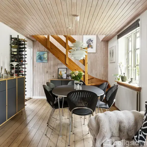 Spisebord med sorte stole står under en trætrappe i et lyst rum. Kunst på væggene og planter på en skænk og vindueskarm skaber en hyggelig atmosfære.