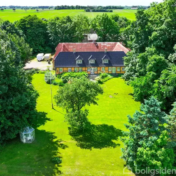 Et spisestuebord med stole står i et rummeligt, gult rum med dekorative tæpper, rødbrune gardiner og antikke møbler. Værelset er fyldt med dagslys fra flere vinduer.