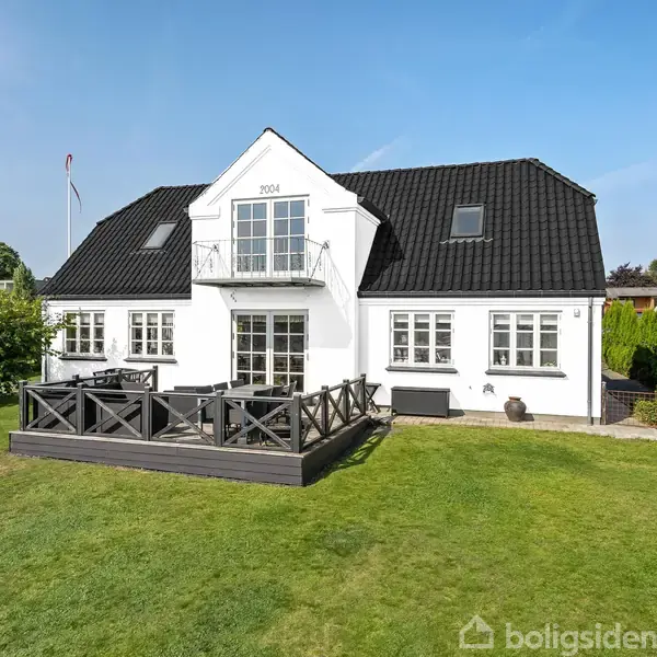 Et hvidt hus med sort tag står stille i en velplejet have med græsplæne. Huset har en terrasse foran, omgivet af buske og træer. 1904 vises på husets facade.