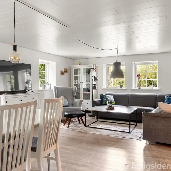 Stue med grå sofa placeret langs væggen, sofabord i midten, TV på skænk, spisebord med stole i forgrunden. Store vinduer slipper lys ind, og lamper hænger fra loftet.