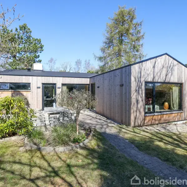Et moderne træhus står stille i en solrig have. Det har store vinduer og stenbelægning foran indgangen, omgivet af grønne træer og buske.