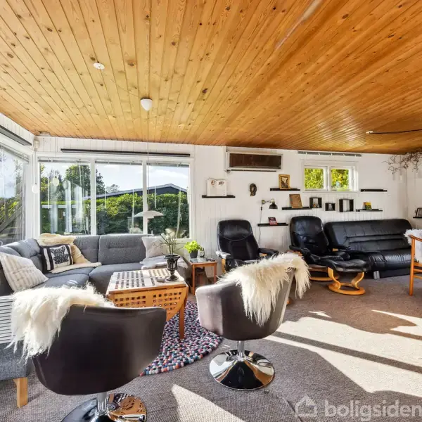 Et lyst stueområde med sofaer og stole dækket af pelse, arrangeret omkring et sofabord. Store vinduer slipper sollys ind, og der er træloft samt boghylder med dekorationer.