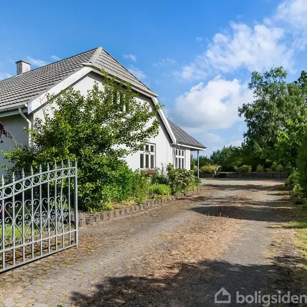 Hus står i solskin omgivet af grønne træer og buske. En bred grusvej løber langs huset, flankeret af en åben smedejernslåge. Blå himmel og lette skyer i baggrunden.
