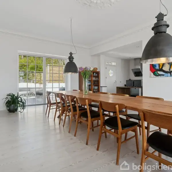 Et langt træbord står med mørke stole omkring det, oplyst af to loftslamper. Rummet er lyst med lyse vægge, et stort vindue og en dør til haven. Der hænger farverige malerier og står planter i hjørnet.