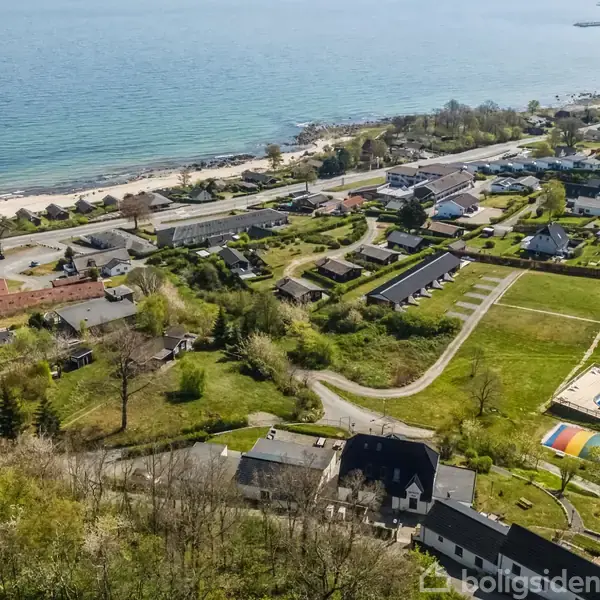 Kystnært boligområde med huse ligger tæt ved stranden. En opblæst legeområde ses nær et hus. Flere grønne områder omgiver bebyggelsen. Havet strækker sig langsomt mod horisonten.