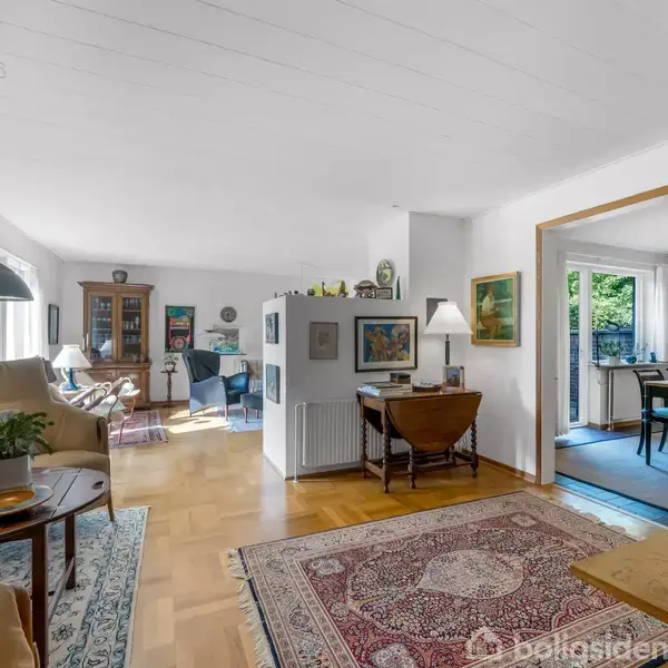Et lyst og rummeligt stueområde med trægulv, sofaer, og tæpper. Væggene er dekoreret med kunst, og en døråbning fører til en spisestue med bord og stole.