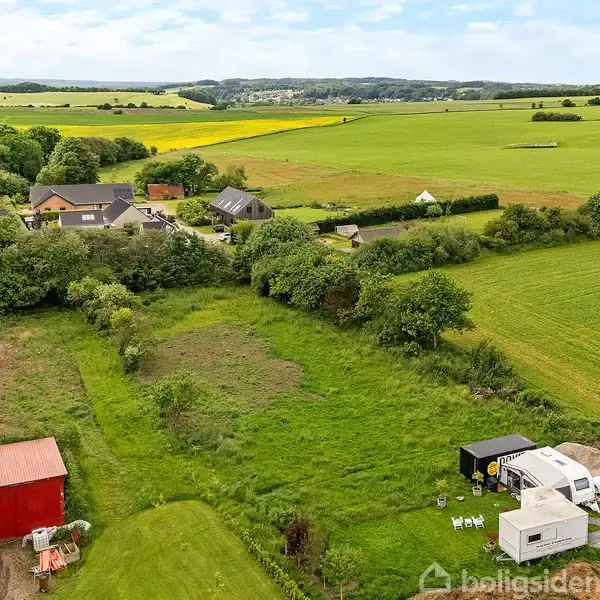 Rød bygning og to trailere står på en grøn mark, omgivet af huse og landområder. Landskabet strækker sig til horisonten med træer og marker.