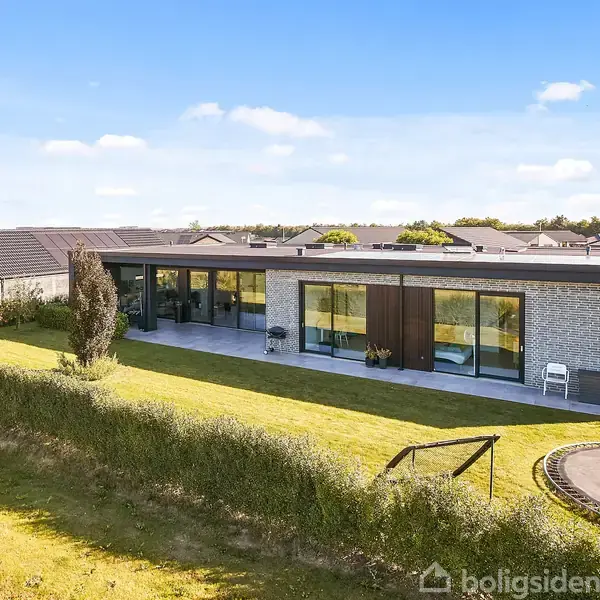 Et moderne hus i ét plan med store glaspartier, placeret i et grønt område med kortklippet græsplæne. Der er en trampolin til højre. Klar blå himmel i baggrunden.
