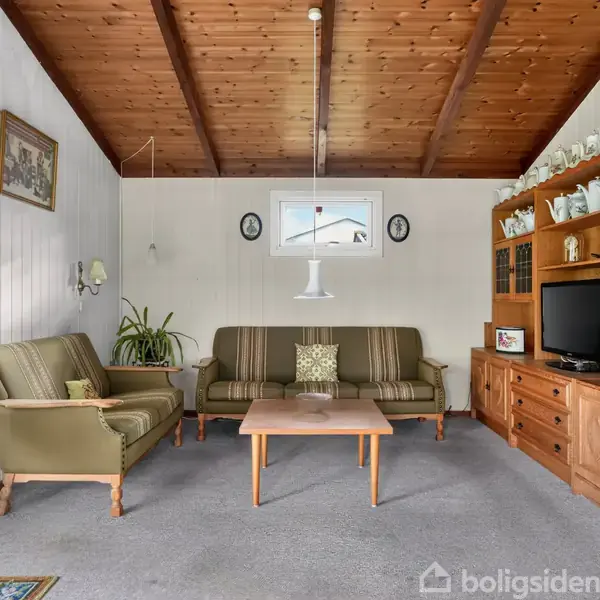 Stue med to grønne sofaer centreret omkring et træsofabord. En lampe hænger over bordet. Tv-bord med porcelænsfigurer og tv. Træloft og lyse vægge. Gardiner ved vinduer.