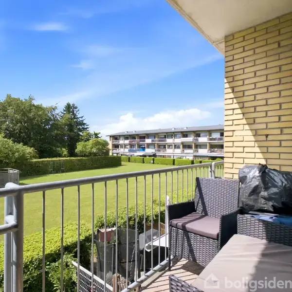 Altan med sort flettet havemøbel, roligt placeret mod en grøn have. Murstensvæg og metalgelænder omfavner rummet, mens en stor bygning ses i baggrunden under en blå himmel.