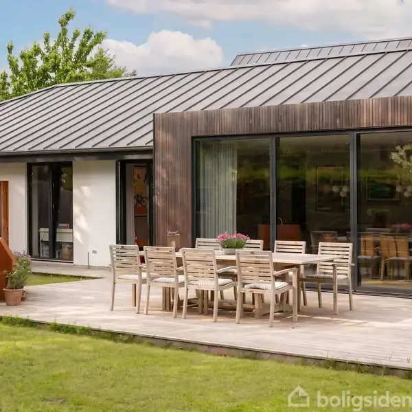 En moderne villa med fladt tag har en rummelig træterrasse. En stor spisebordssæt med stole står på terrassen. Græsplæne omkranser huset, og træer ses i baggrunden. Ingen tekst.