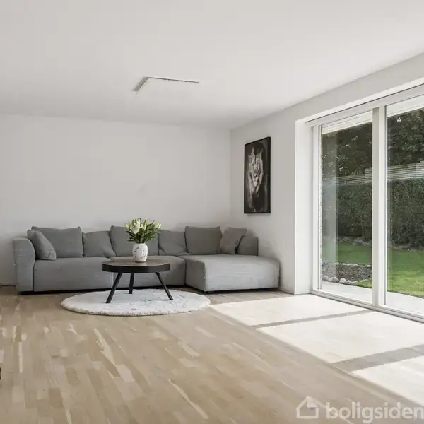 Grå sofa står stille i en lys stue med trægulv. Rundt sofabord er dekoreret med blomster. Store vinduer med havudsigt fylder væggen. Væggene er hvide med enkel kunst.