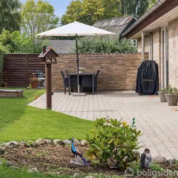 Terrasse med flisebelægning har et sæt havemøbler og en parasol. Græsplæne og planter omkranser området, mens en stensat blomsterbed står i forgrunden. Grænsehæk til venstre.