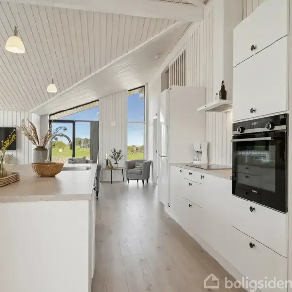 Et moderne køkken med hvide skabe og indbygningsovn står i et lyst rum. Stuen er synlig i baggrunden med store vinduer, der lukker naturligt lys ind.