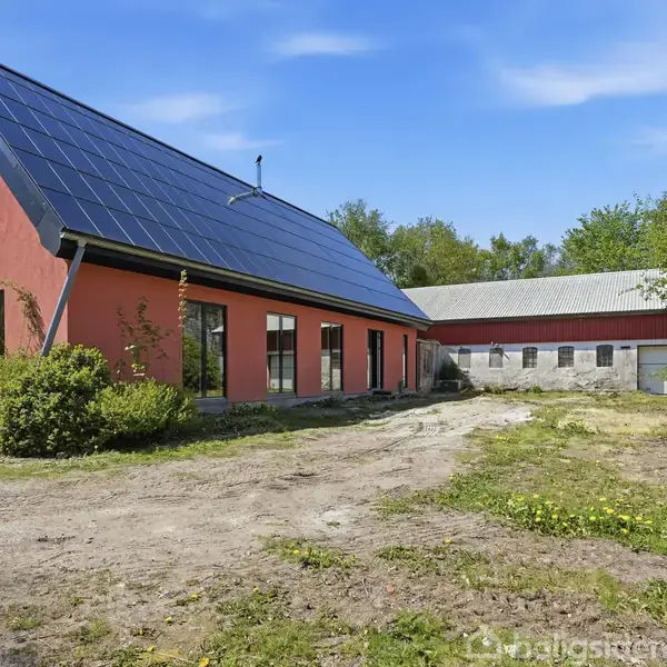 Et moderne hus med røde facader og solpaneler på taget. Huset står på en grusbelagt gårdsplads omgivet af grønne buske og træer, med en stor lade i baggrunden.