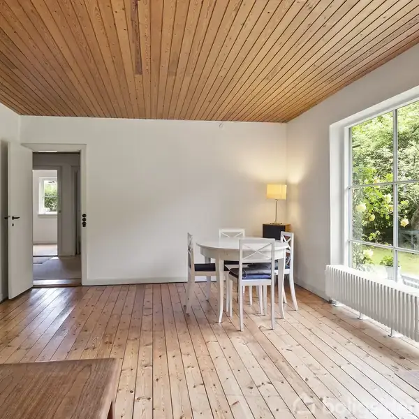 Spisebord med stole står ved et stort vindue i en stue med trægulv og træloft. En lampe lyser ved bordet, og haven kan ses udenfor gennem vinduet.