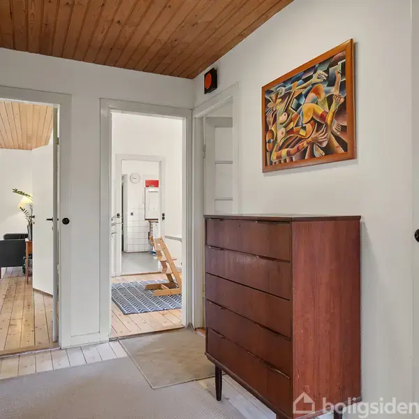 Kommode står stille i en husgang med træloft og tre døre. Forgrunden har en farverig kunstplakat på væggen. Indgangene fører til stuer med trægulve og en flisebeklædt badeværelse.