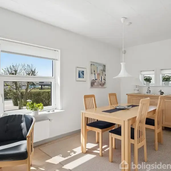 En lys spisestue med et træbord og fire stole, en loftlampe hænger over bordet. Store vinduer giver udsigt til en have, og en sort lænestol står ved siden af bordet.